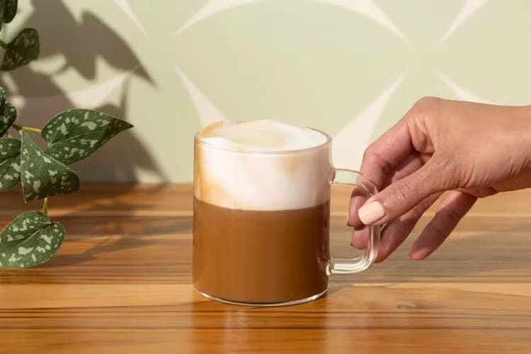 A hand holding a Starbucks Caffè Misto in a translucent mug, on a wooden surface