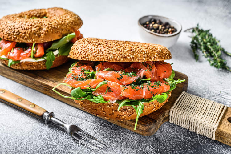 Two smoked salmon bagels on a wooden serving board, in between a fork and pepper corns