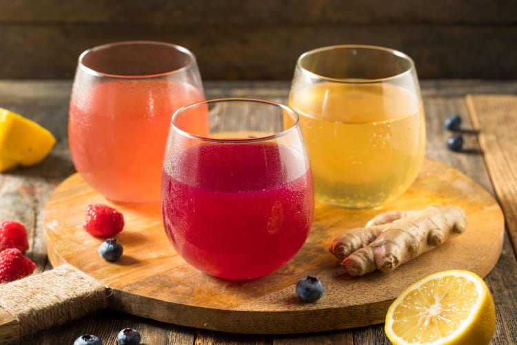 Orange, red and yellow kombucha drinks on a wooden table surrounded by fresh fruit.