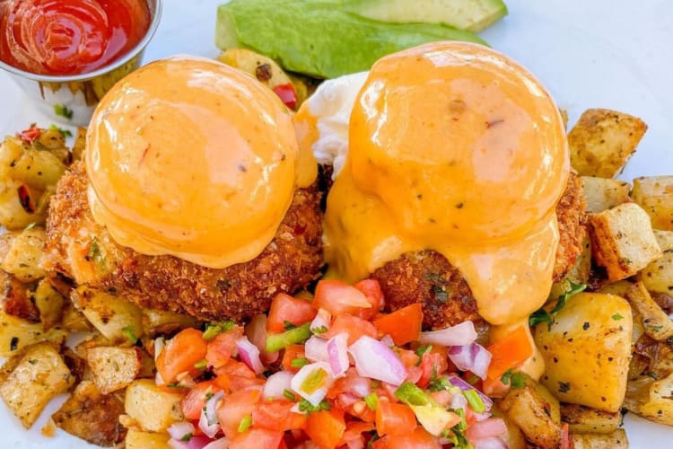 Crab cakes under poached eggs, topped with sauce, served with avocado and potatoes