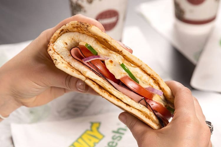 Hands holding a Subway flatbread sandwich with eggs and vegetables