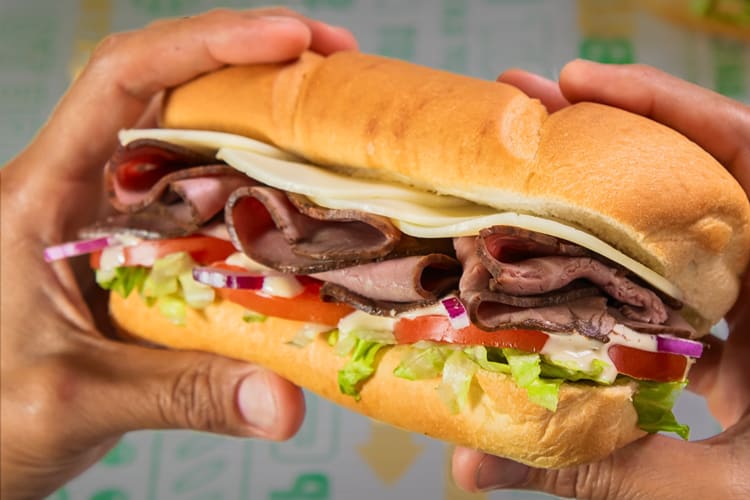 Hands holding a Subway sub with meat and vegetables