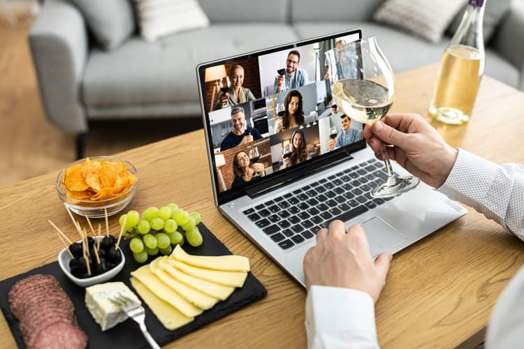A person's hands holding a wine glass in front of a laptop, next to snacks, during a virtual wine tasting