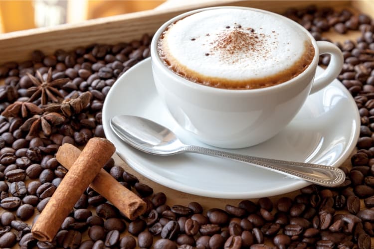 A cappuccino next to coffee beans and two sticks of cinnamon