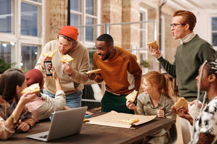 a group of people laugh and joke together eating pizza in a workplace