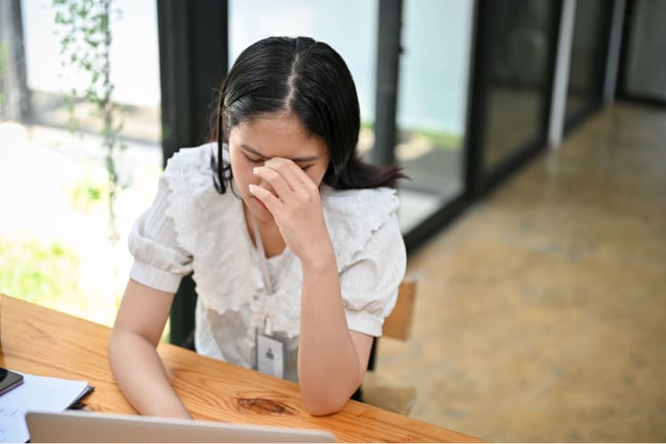 a woman sits at her desk with her head in her hands, clearly unhappy at work