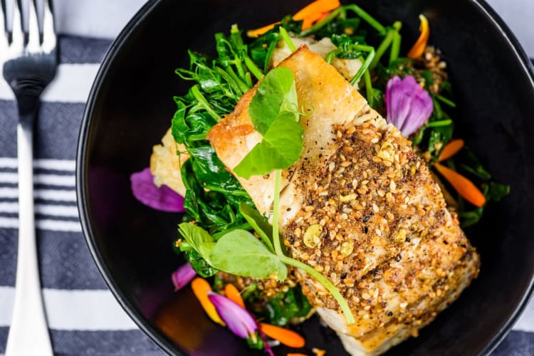 A filet of barramundi fish baked with sesame seeds, presented over a colourful salad.