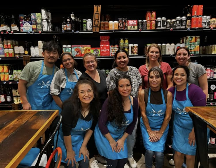 Participants in Classpop! aprons pose together in front of shelves of craft beer in a mixology team building activity in Boston.