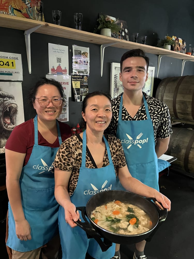 Three participants wearing Classpop! aprons pose with a large pan of prepared food during a cooking class in Phoenix.
