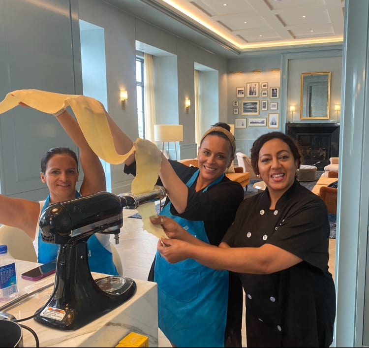 Three people hold a long sheet of pasta dough beside a stand mixer fitted with a pasta roller in a cooking class in Phoenix.