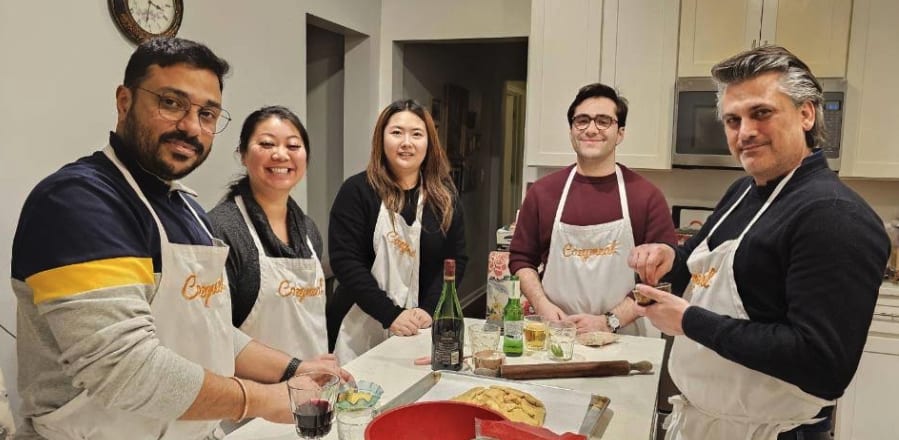 Five participants wearing Cozymeal aprons gather around a kitchen counter with pasta dough and rolling pins for a  cooking class in NYC.