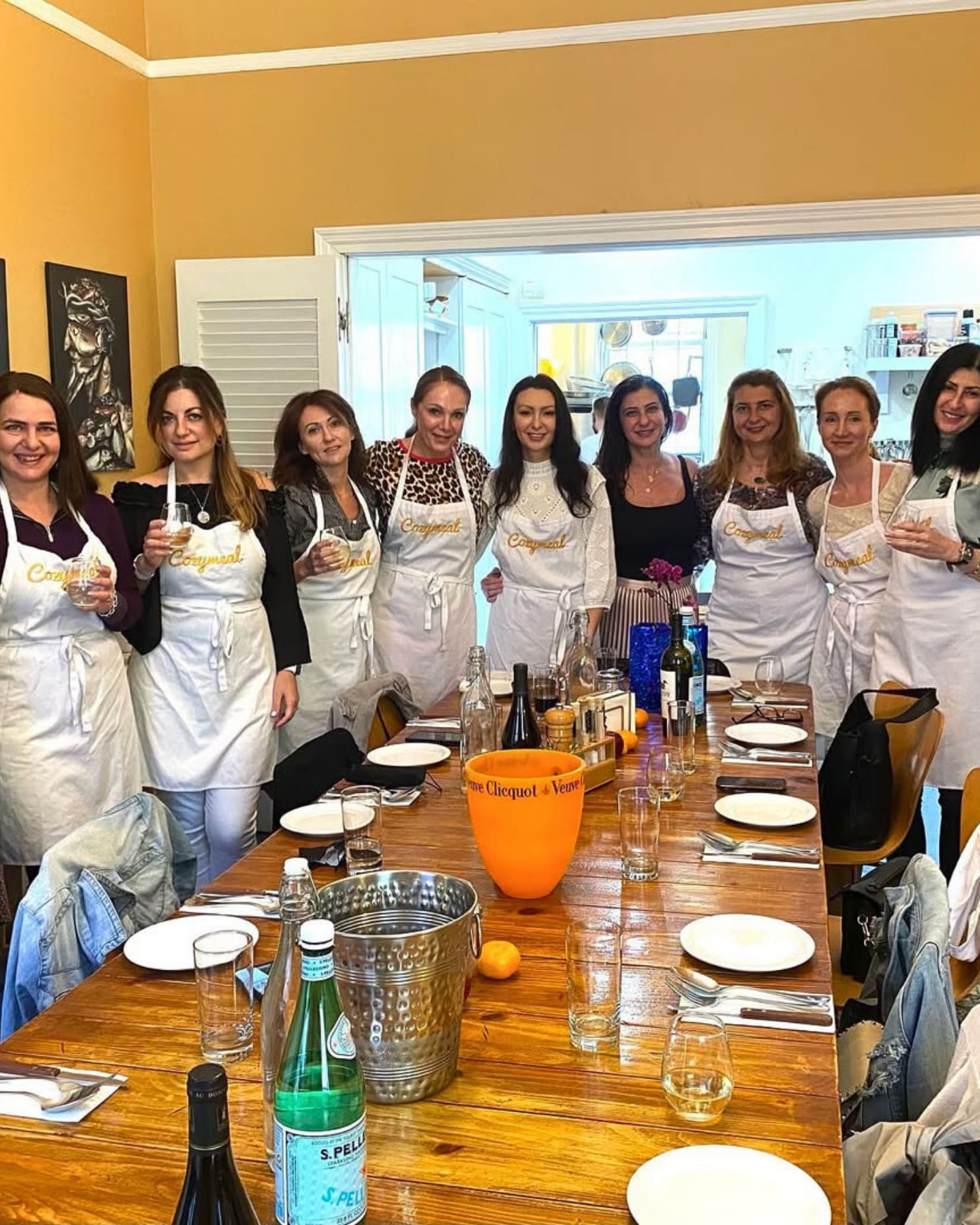 Group wearing white Cozymeal aprons poses beside a dining table set with plates, glasses and bottles.