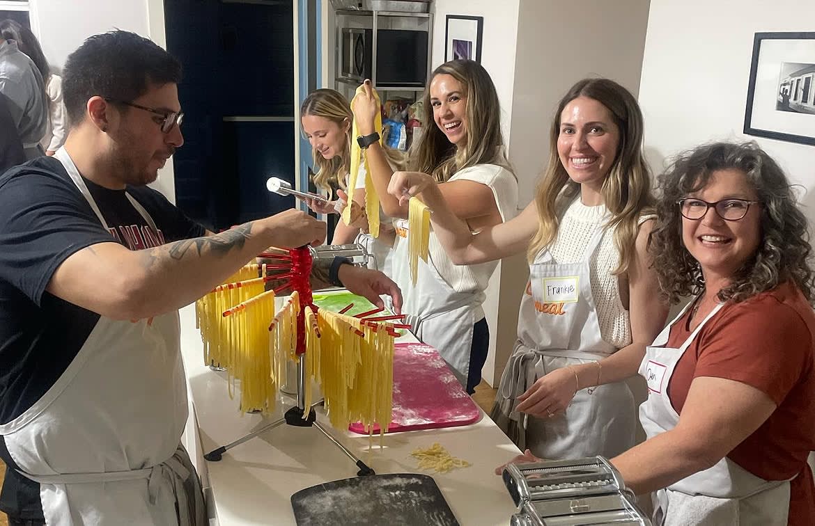 Participants wearing Cozymeal aprons make fresh pasta together around a kitchen counter in a team building activity in San Francisco.