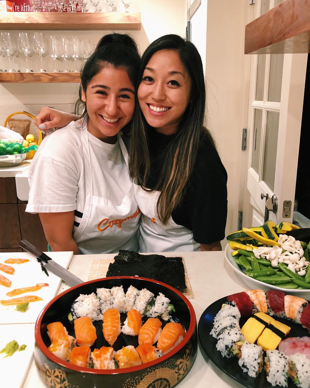 Two guests wearing Cozymeal aprons in a sushi making class in San Francisco pose beside plates of sushi, with rice, seaweed and vegetables on the counter.
