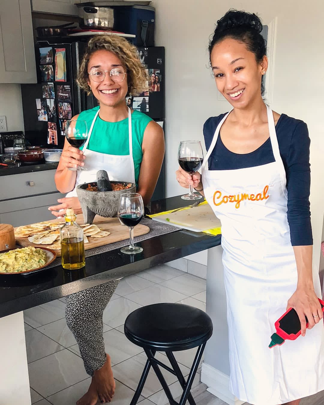 Two participants wearing Cozymeal aprons hold glasses of red wine beside a kitchen counter with prepared food in a Mediterranean cooking class in Los Angeles.