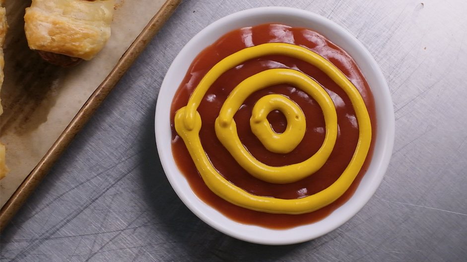 Mummy Dogs Recipe: Using either the nozzle of the mustard bottle or a pastry bag fitted with a small round tip, pipe concentric circles on top of the ketchup.