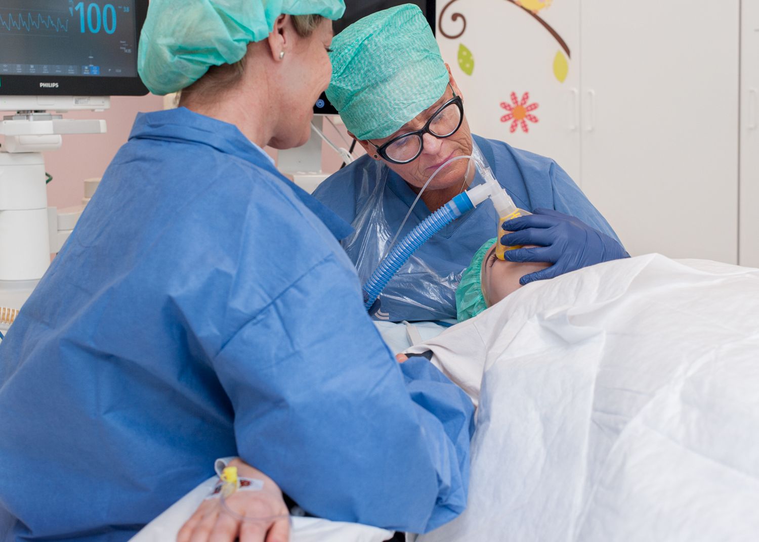 En patient ligger på ett operationbord och andas i en syretratt. En anhörig sitter bredvid operationsbordet.