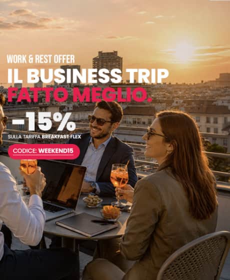 Un banner pubblicitario ambientato su una terrazza panoramica a Milano al tramonto, con vista sul Duomo e sullo skyline della città. In primo piano, alcune persone in abbigliamento business-casual chiacchierano a un tavolo sorseggiando dei drink. Sull'immagine campeggia un testo promozionale in bianco e rosa che offre uno sconto del 15% sulla tariffa "Breakfast Flex" per i soggiorni nel weekend, utilizzando il codice "WEEKEND15" sul sito bbhotels.com.
