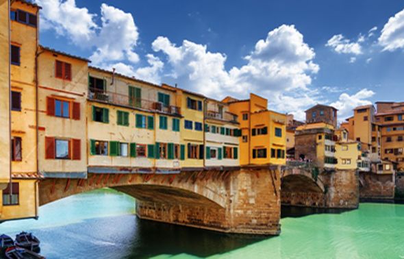The historic Ponte Vecchio in Florence, showcasing its iconic colorful medieval shops built over the Arno River. The turquoise water reflects the yellow and orange buildings under a bright blue sky filled with fluffy white clouds.