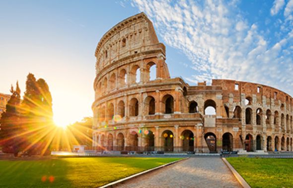 A wide-angle view of the Colosseum in Rome under a bright blue sky with scattered clouds. The morning sun bursts from behind the trees on the left, casting a vibrant lens flare across the ancient Roman amphitheater and the green grass in the foreground.