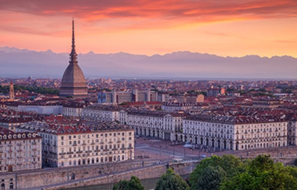 An elevated panoramic view of the city of Turin at sunset. The prominent dome and spire of the Mole Antonelliana stand out against the city skyline, with the majestic, snow-capped Alps visible in the background under a vibrant pink and orange sky.