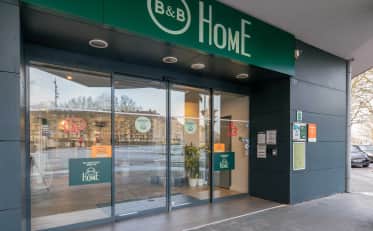 Moderner Eingang des B&B HOME Hotels mit Glasschiebetüren und grünem Schild