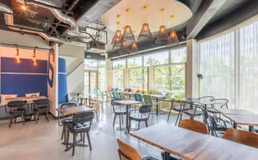 Restaurante de hotel moderno com design industrial, mesas de madeira e grandes janelas iluminadas.