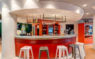 Moderne Hotelbar im B&B HOME mit leuchtend roter Theke und stilvollen Metallhockern für eine entspannte Atmosphäre