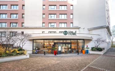 Außenfassade und Eingang des modernen B&B HOME Hotels mit gläserner Lobby und Sitzbereich im Freien