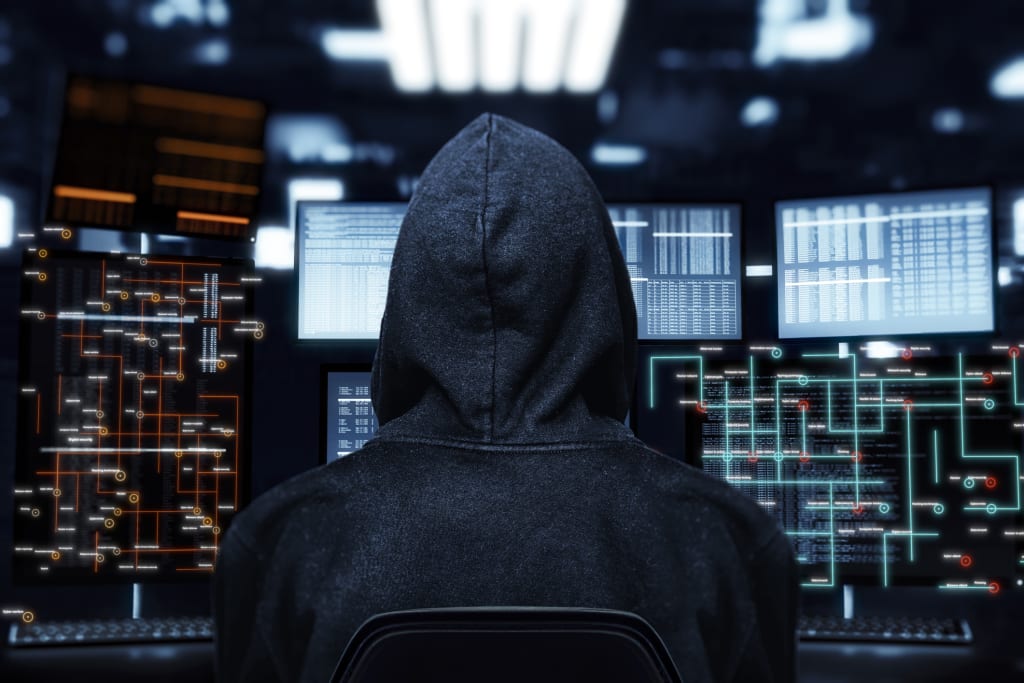 Dark hooded figure sitting in front of several futuristic screens
