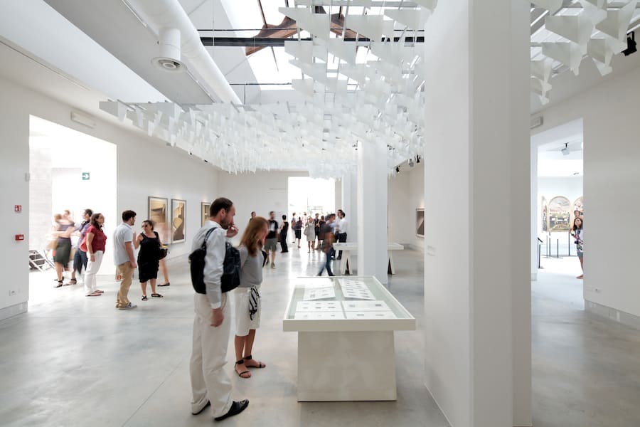 Visitors walking through a bright museum gallery past artworks and a display case
