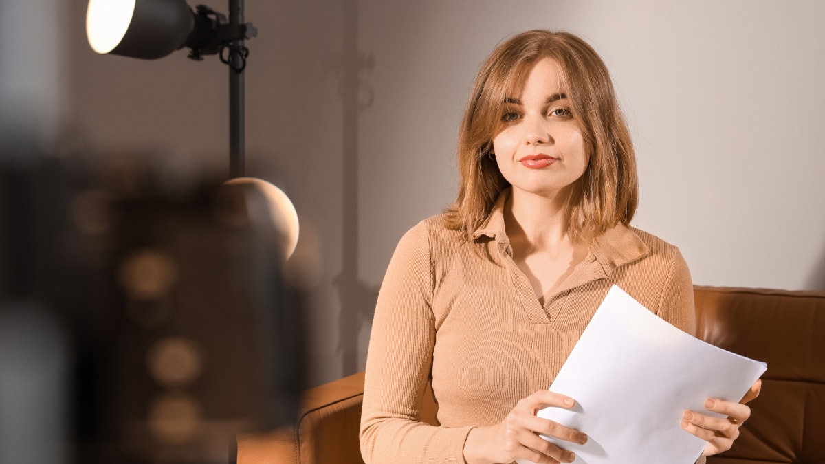 An actor sitting under a studio lamp, holding her sides and looking straight at the camera