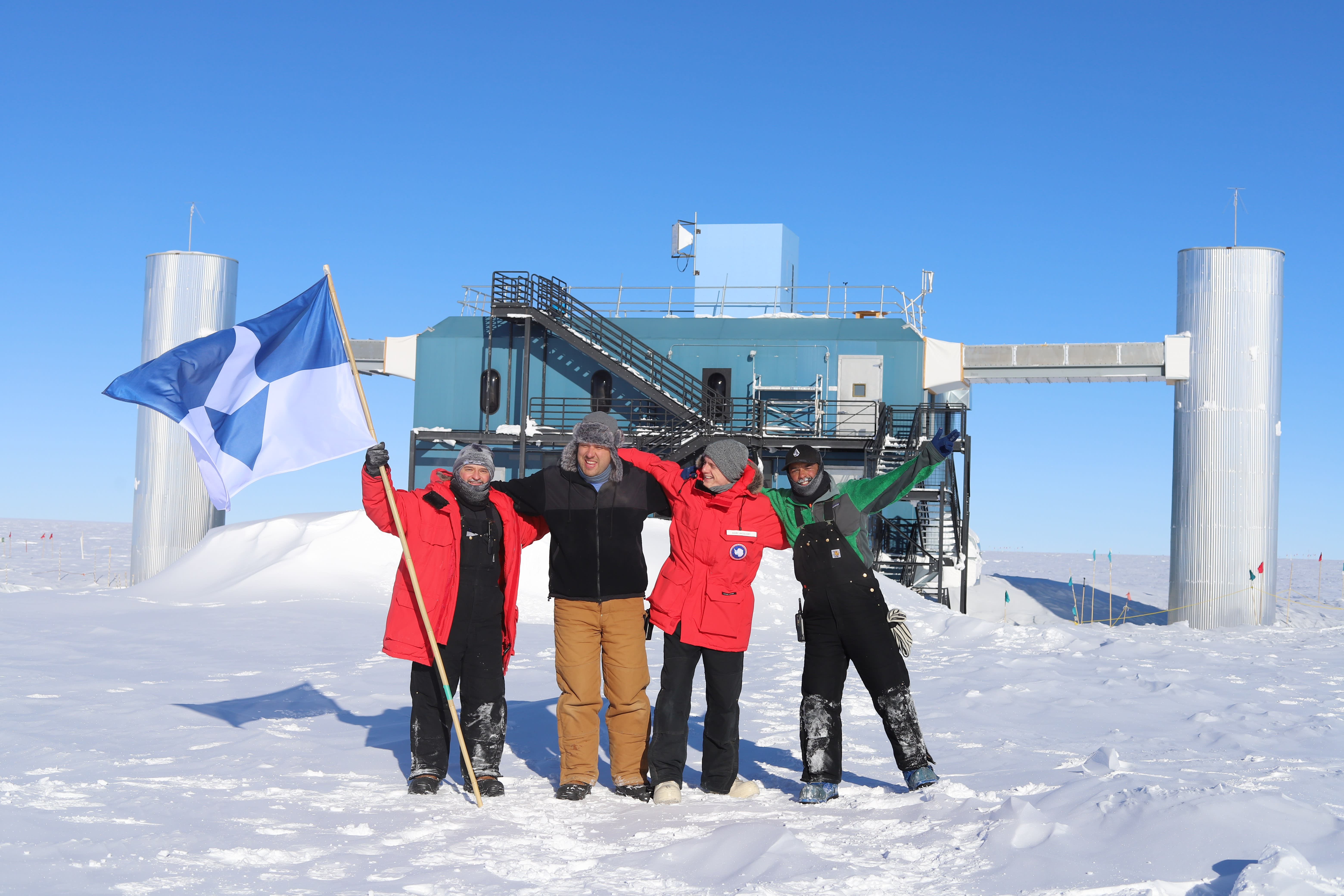 Group photo of four winterovers standing in front of the IceCube Lab under blue sky.