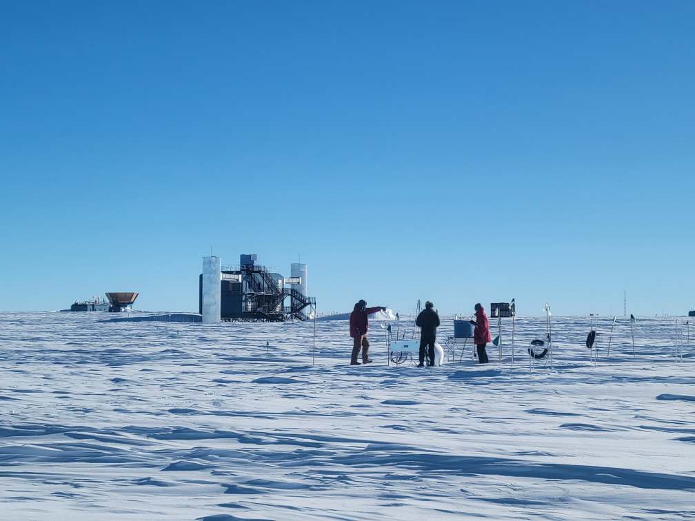 Group working outside on the South Pole ice under clear skies with IceCube Lab in background.