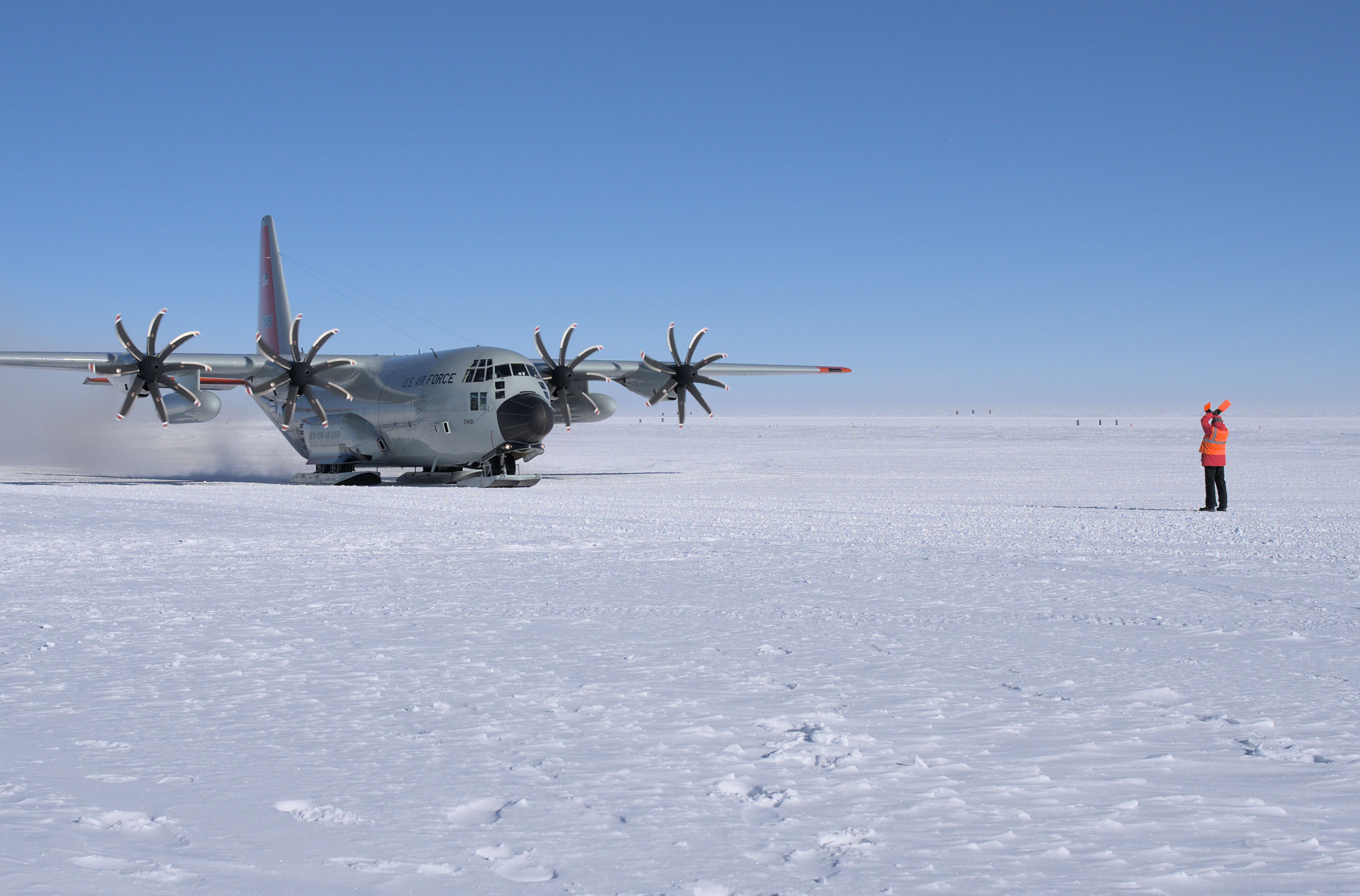 Winterover in red parka out on ice marshalling an LC-130 plane.