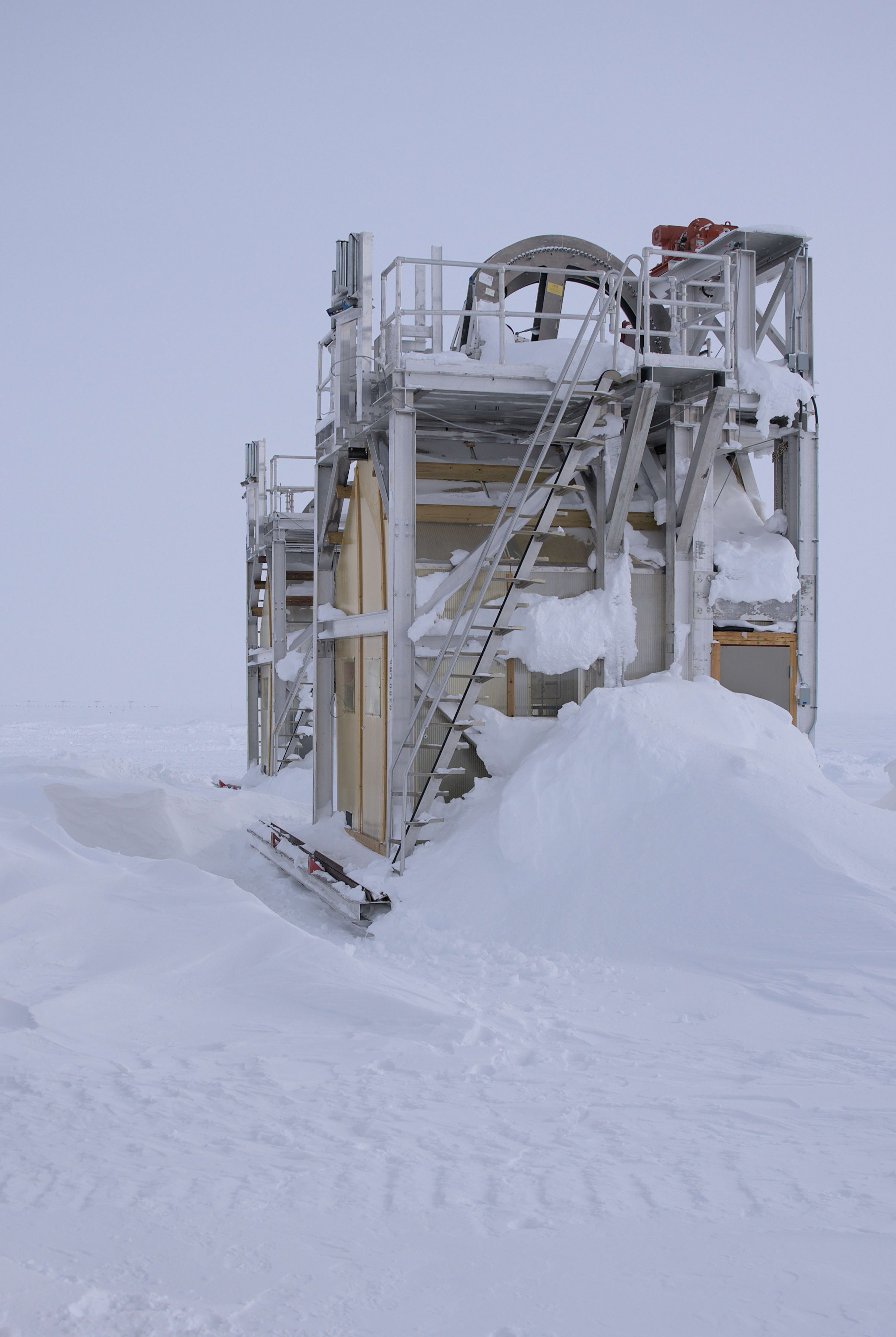 Drill tower covered in windswept snow under cloudy sky.