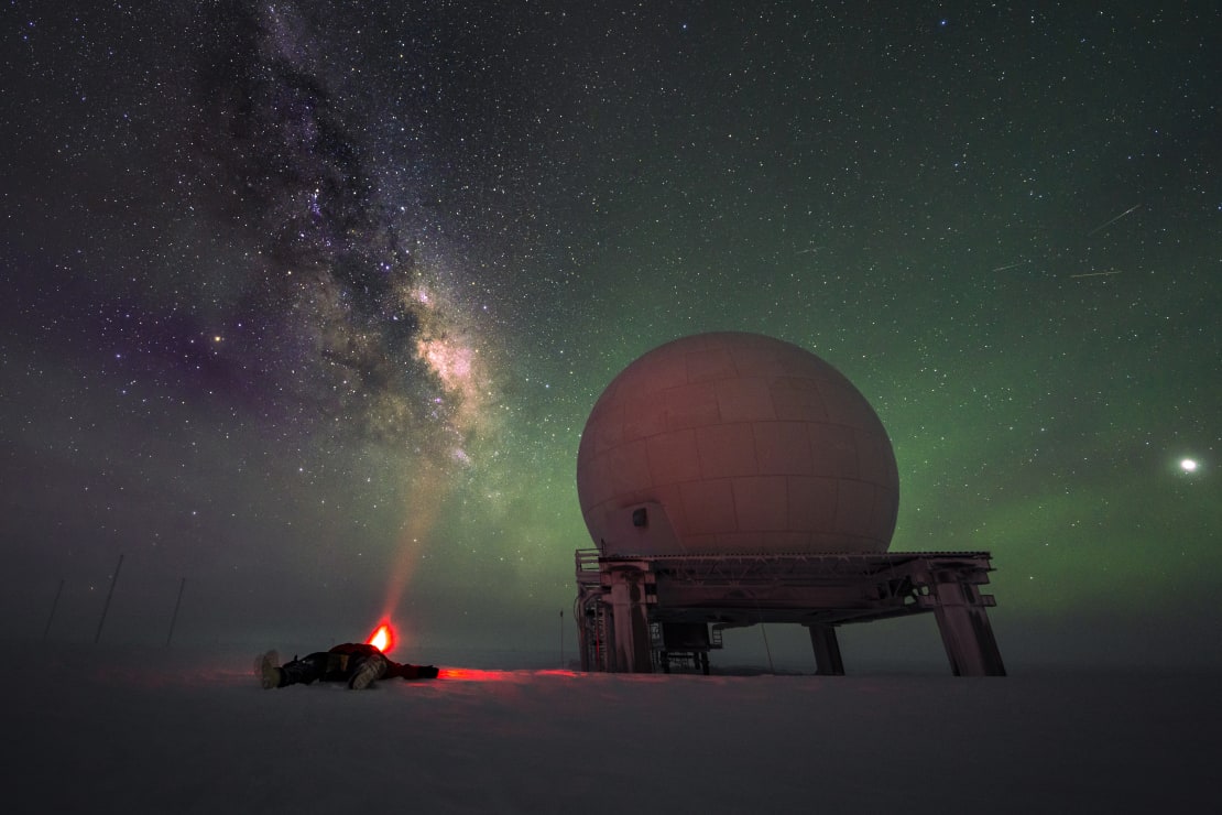 Person lying on ground near satellite dome, with clear view of Milky Way in sky above.