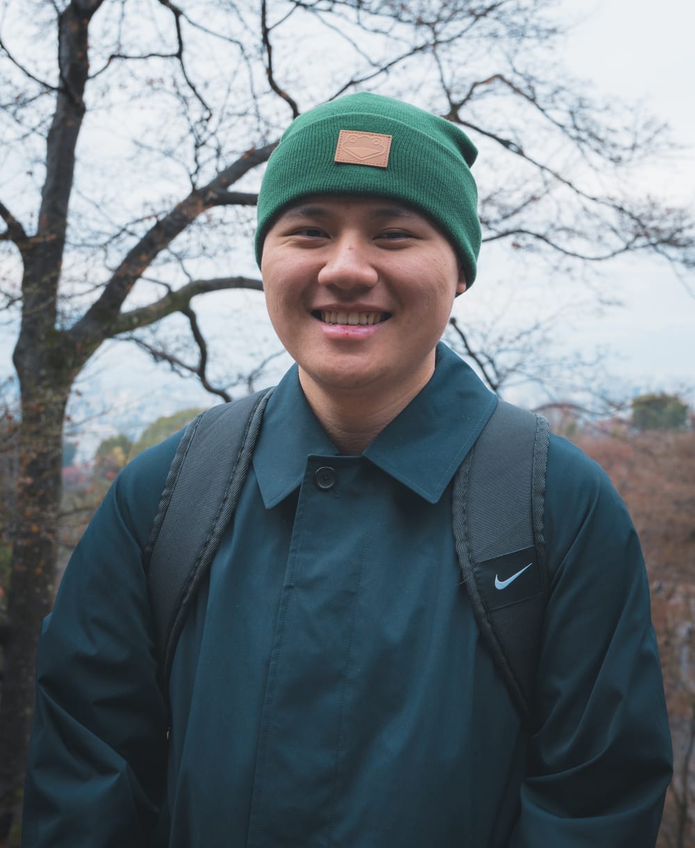 A headshot of a man wearing a beanie outside in front of a tree.