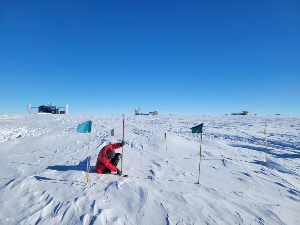 Person taking snow measurements on the South Pole ice under clear skies with IceCube Lab in distant background.