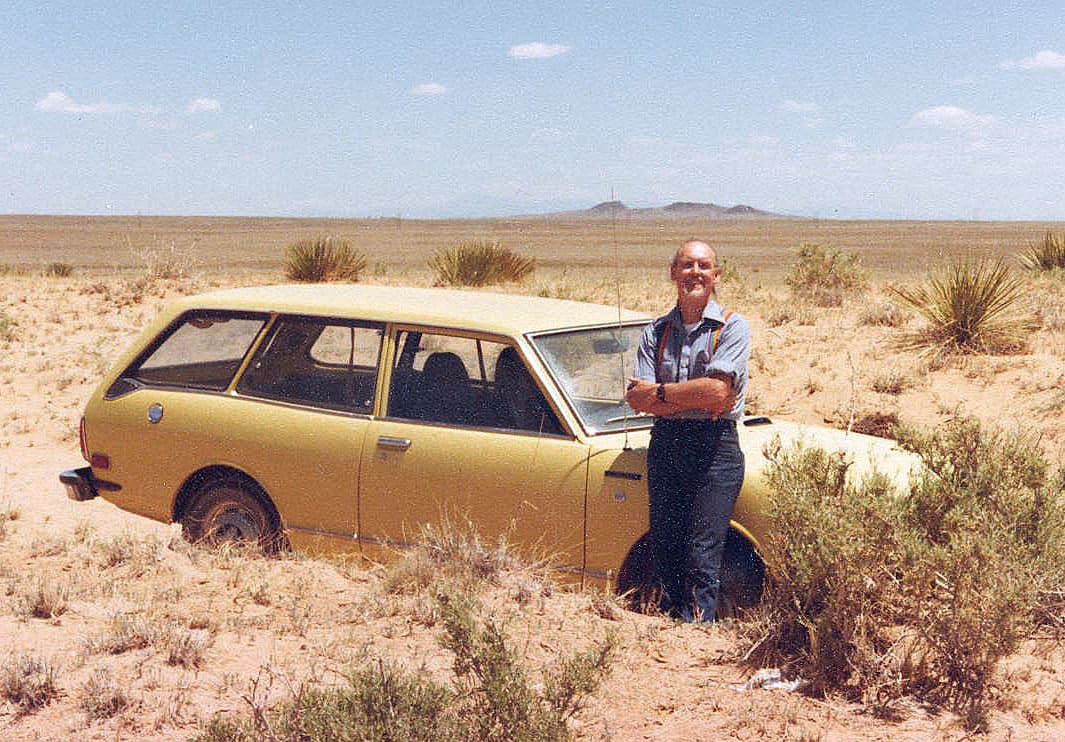 A man wearing rainbow suspenders leaning against a yellow car in a desert landscape.