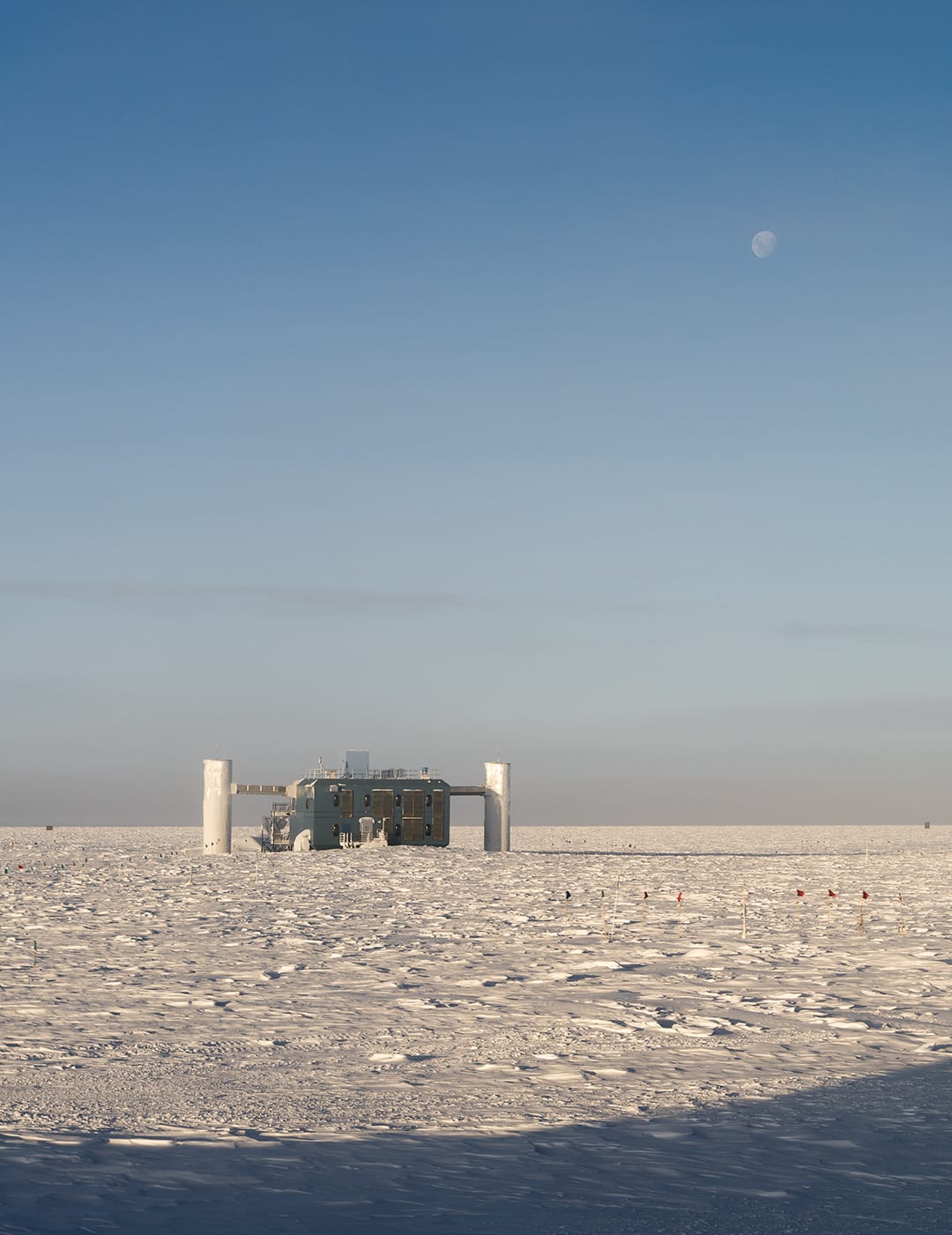 The IceCube Lab sitting in low, bright sunshine with moon high overhead.