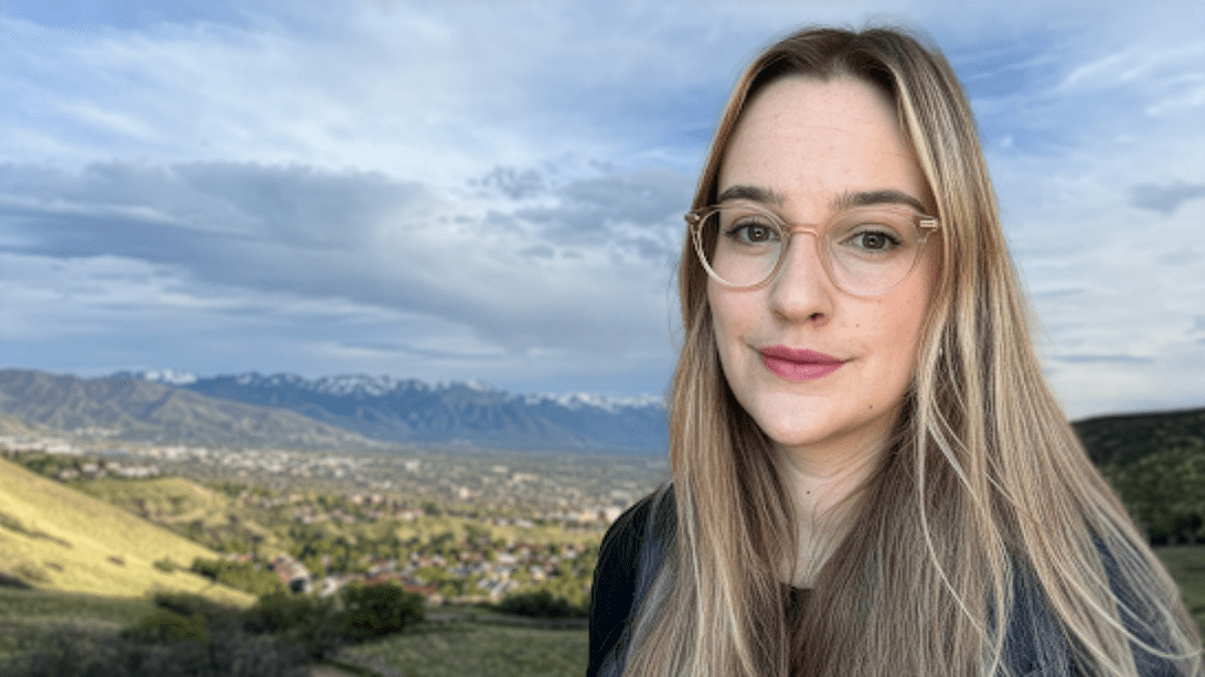 A girl with glasses and blonde hair in front of a backdrop of mountains.