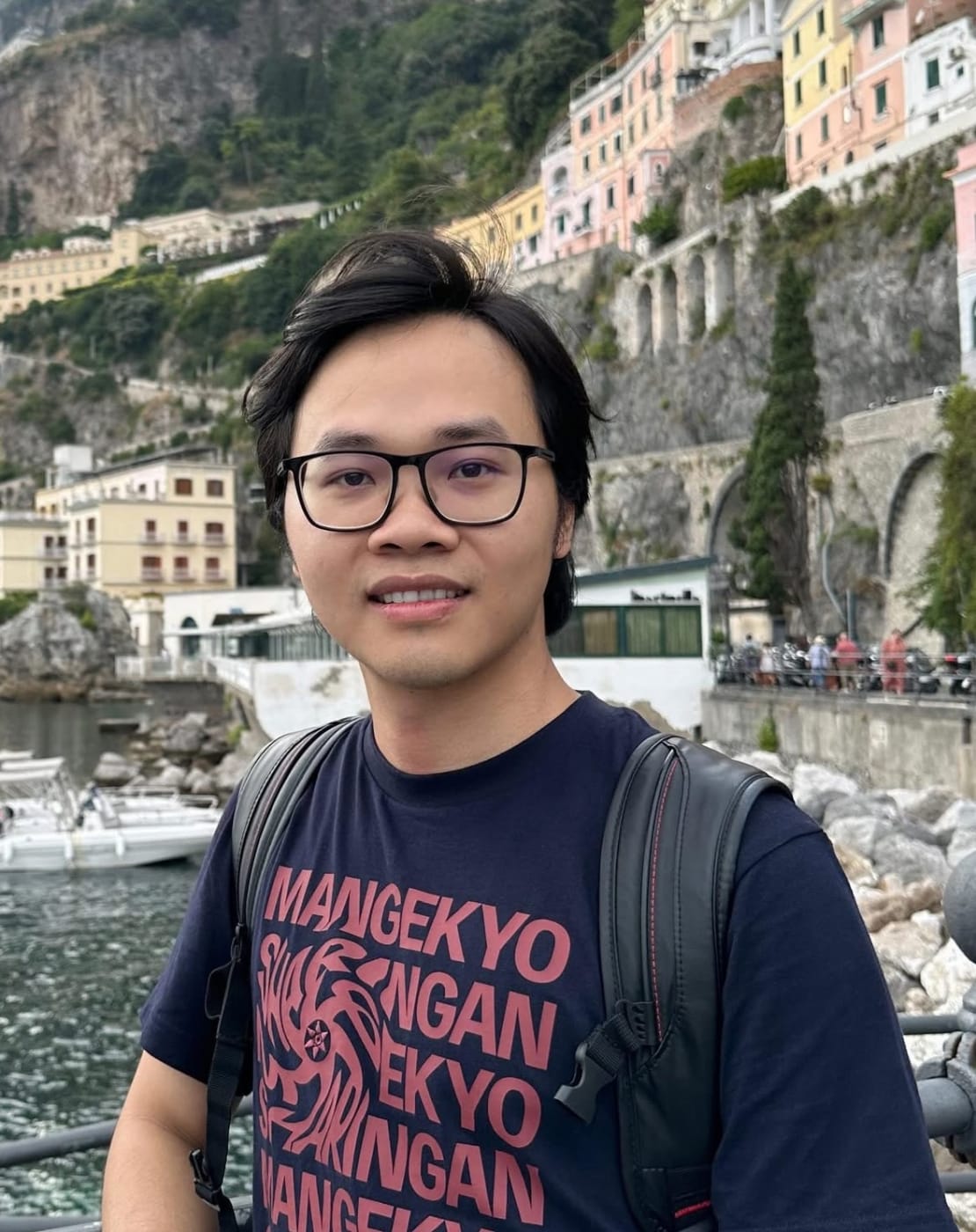 A person with glasses posing in front of buildings on a cliff.