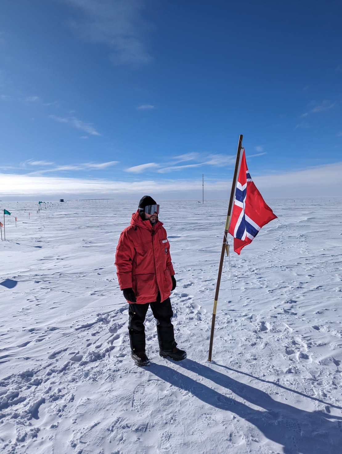 Winterover in red parka standing next to Norwegian flag out on the ice on sunny day with blue sky.