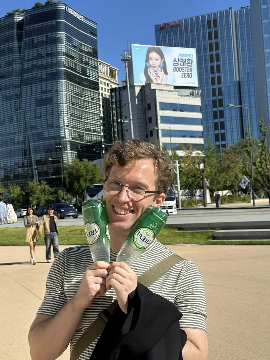 A man with glasses holding two bottles up to their face in front of tall buildings.
