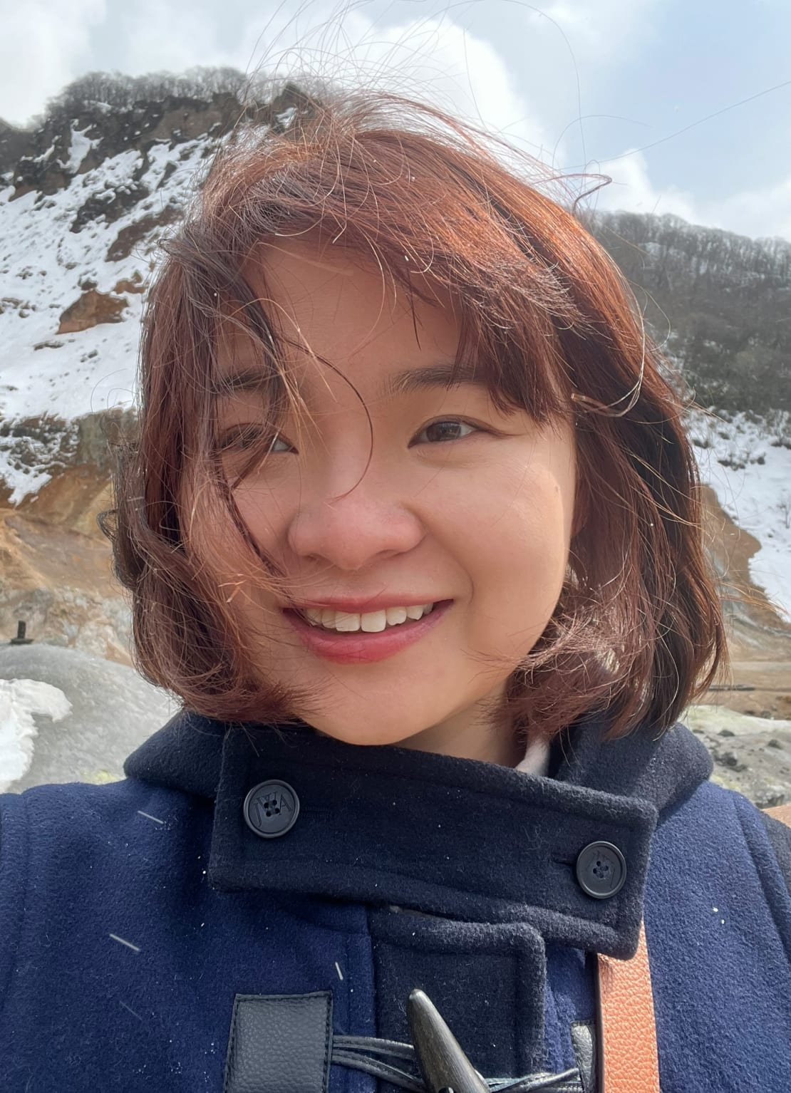 A woman wearing a blue pea coat smiling in front of a snowy mountain.