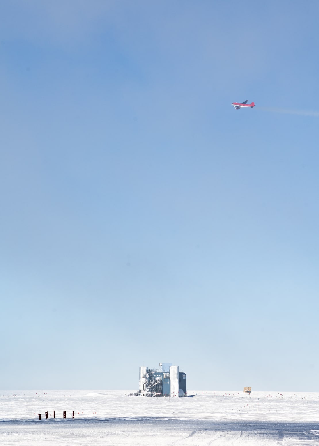 A red airplane high above the IceCube Lab, with clear blue skies.