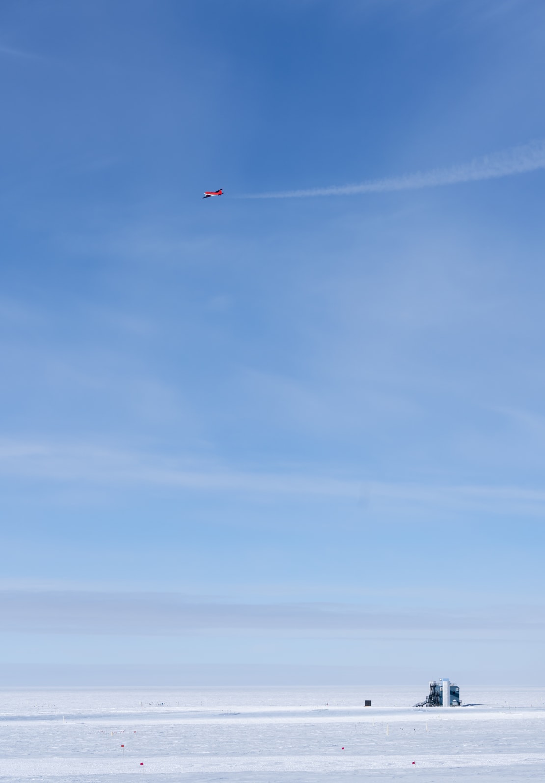 A plane with contrails high in a blue sky over the IceCube Lab, appearing small in lower right.