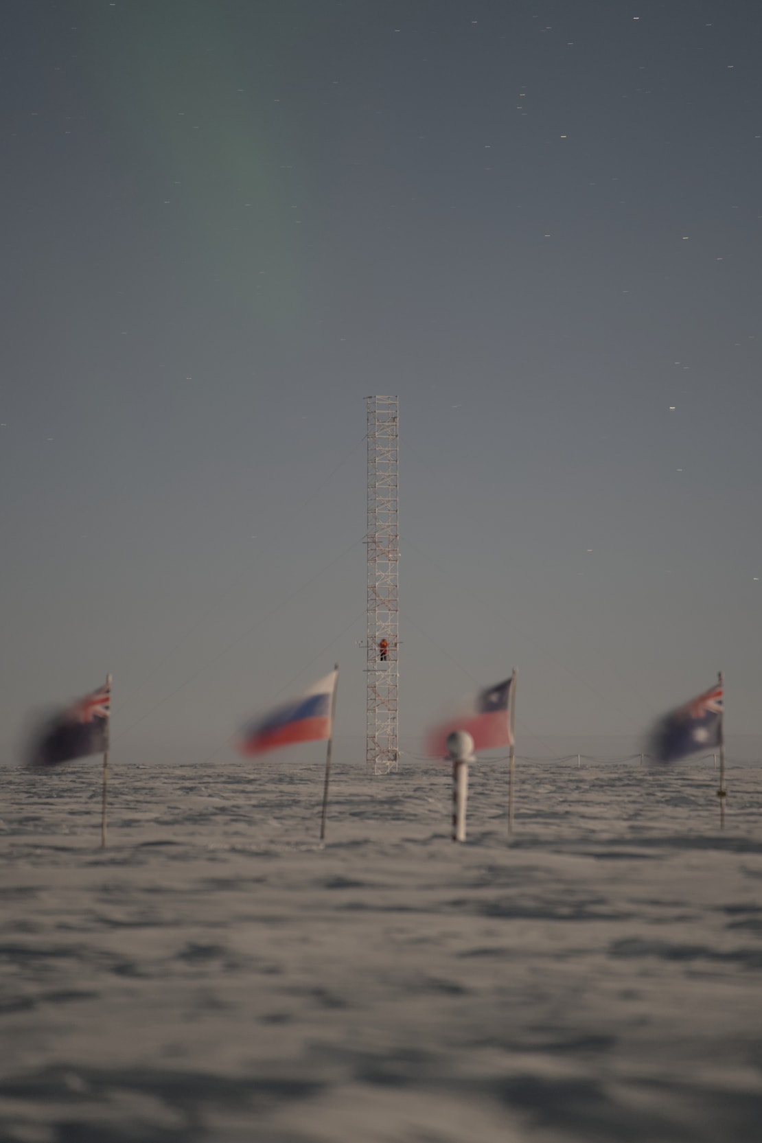 Flags flapping in wind at ceremonial South Pole, with a tall tower structure in distance behind them.