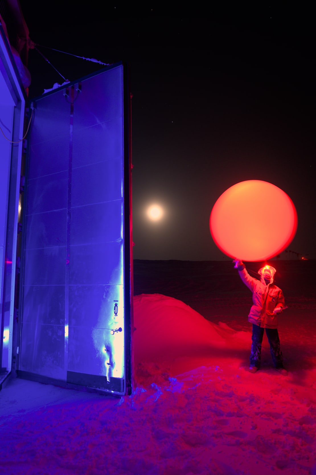 Winterover in parka holding up a weather balloon outside the launch building, lit in red.